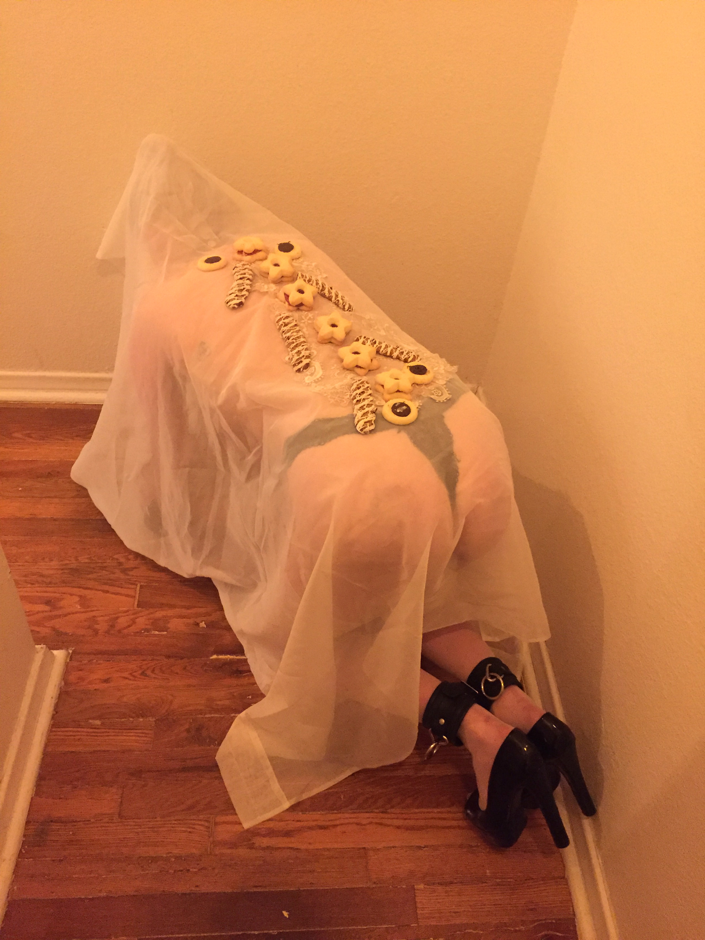 Ariel view of a girl on all fours with a sheer sheet over her and cookies on her back in a narrow hallway in warm light